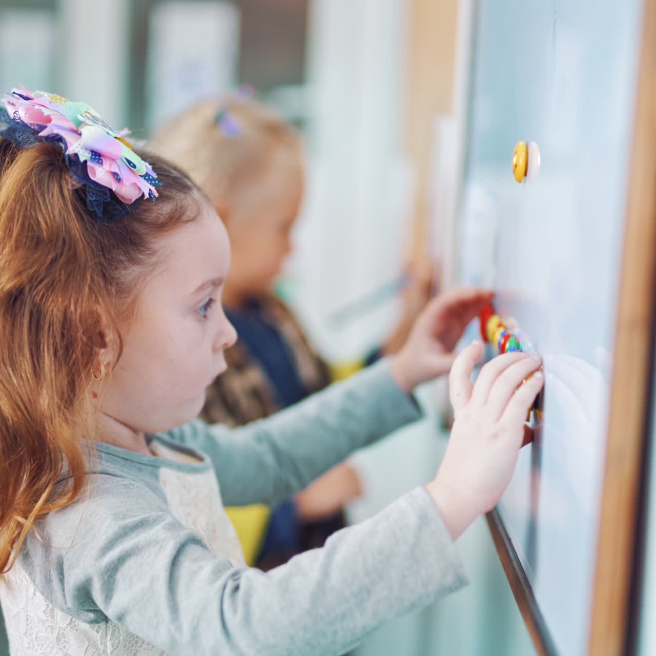 Little girls near the board with magnets in primary school. Children playing with colorful magnets on white board on the background of classroom.