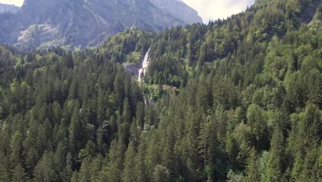 Ein-Drohnenflug-über-Einen-üppigen-Grünen-Alpenwald-In-Richtung-Eines-Kaskadierenden-Wasserfalls-Aus-Gletscherschmelze,-Der-über-Eine-Klippe-Stürzt