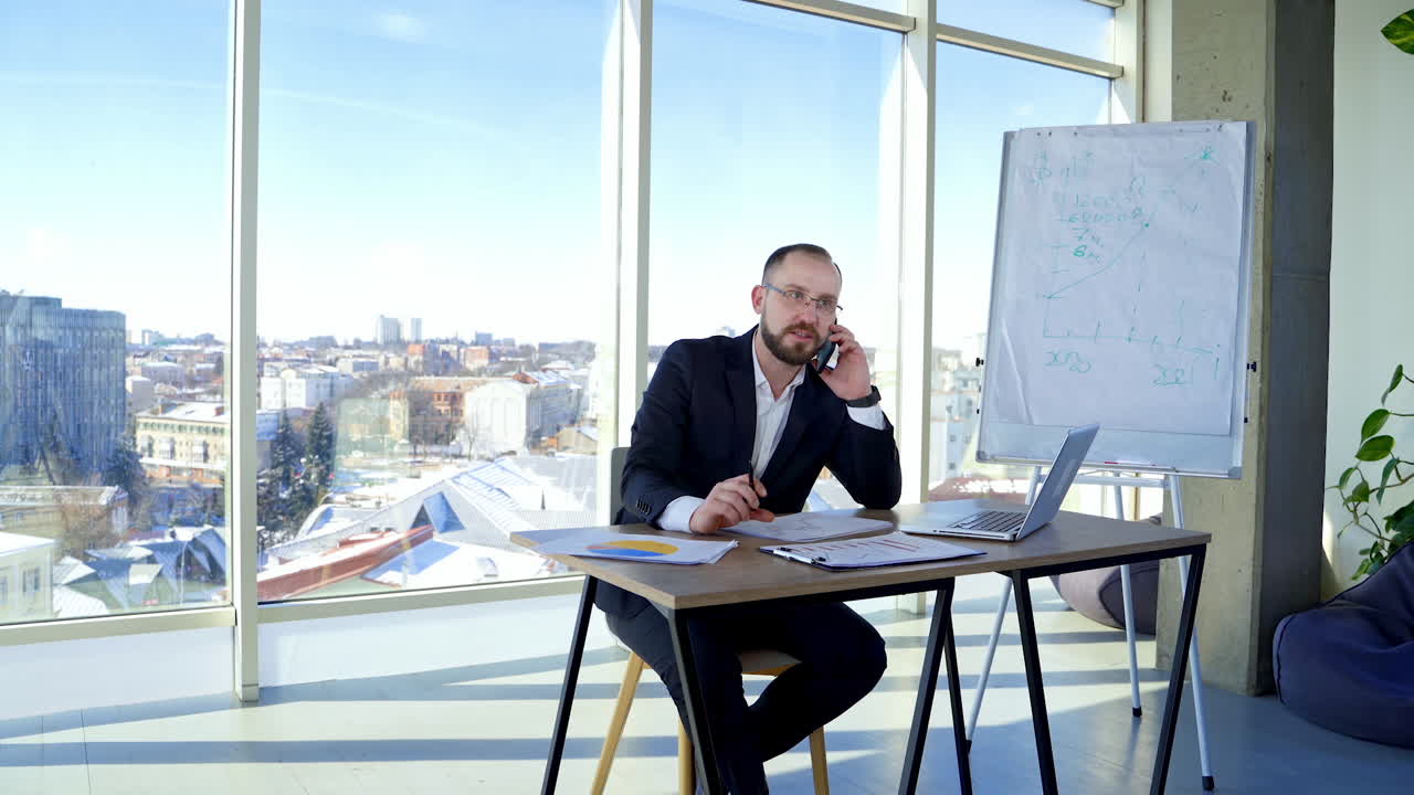 Professional entrepreneur working at office centre. Businessman in suit sitting at his workplace and talking the phone on large windows background.