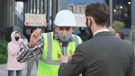 periodista o corresponsal caucásico con máscara protectora en una entrevista con un constructor en una protesta contra el covid 19