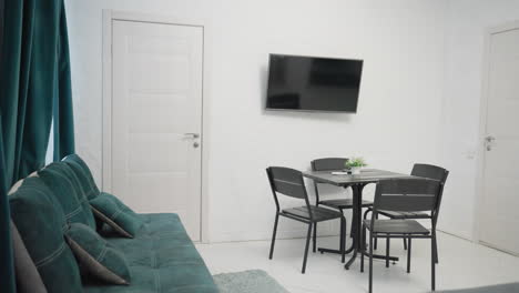modern apartment living area with green velvet sofa, black dining table, mounted tv on white wall, and small potted plant on table. clean white interior with teal curtains and large window
