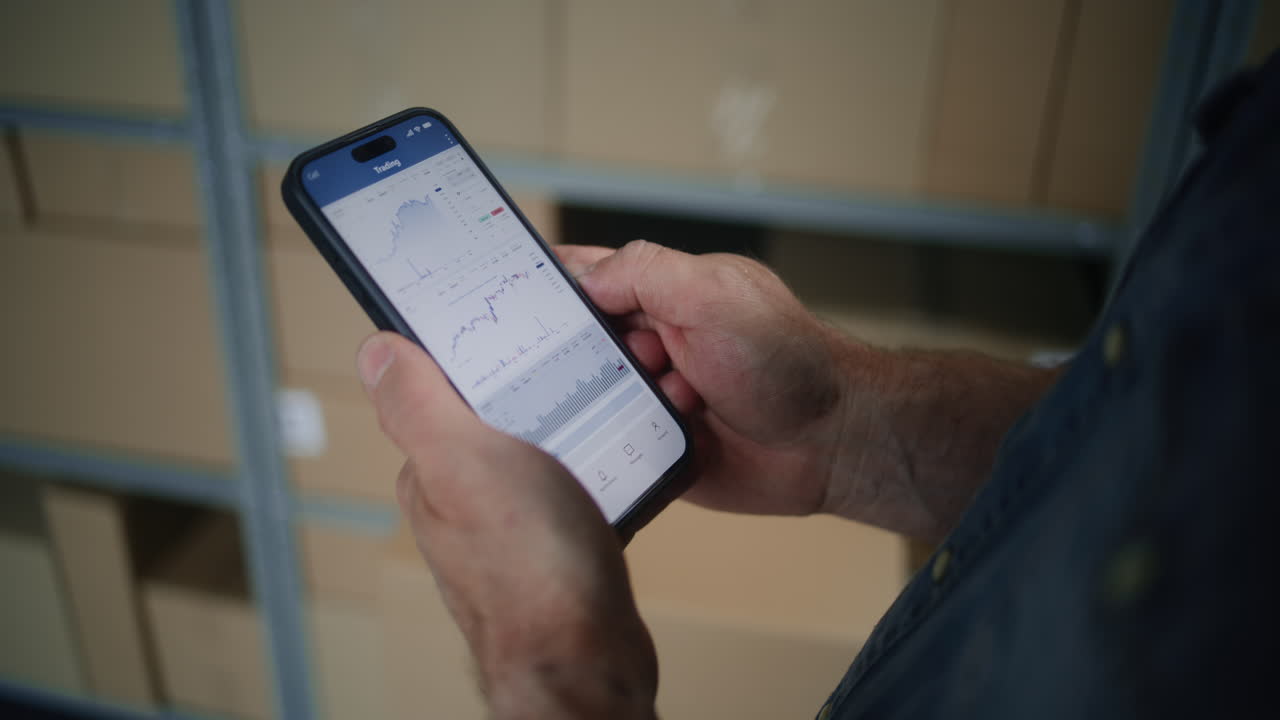 Worker Monitoring Real-Time Stocks, Exchange Market Charts Using Smartphone. Man Making Cryptocurrency Investments During Working Day in Warehouse, Combining Job with Online Trading. Close Up Shot.