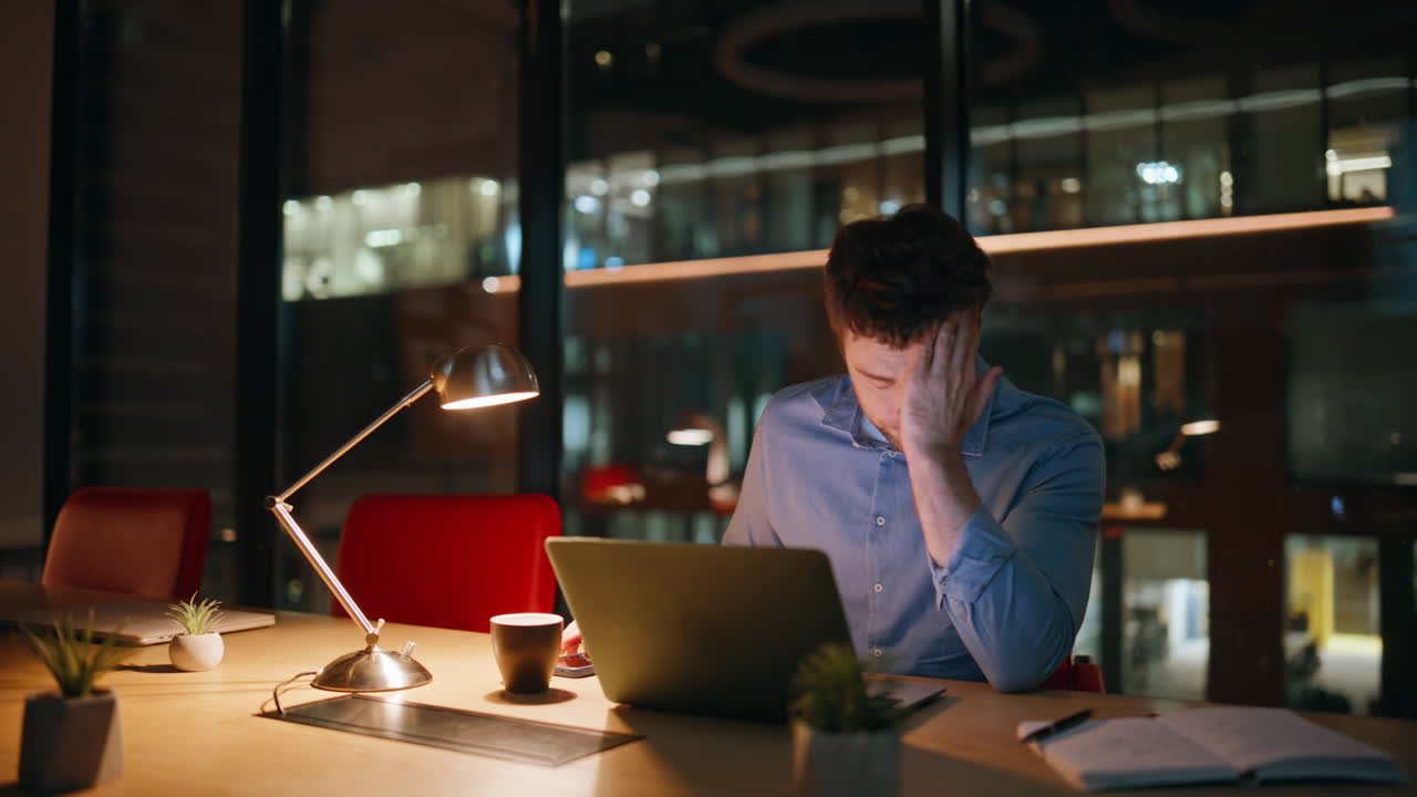 Overworked employee talking cellphone looking laptop at workspace late evening