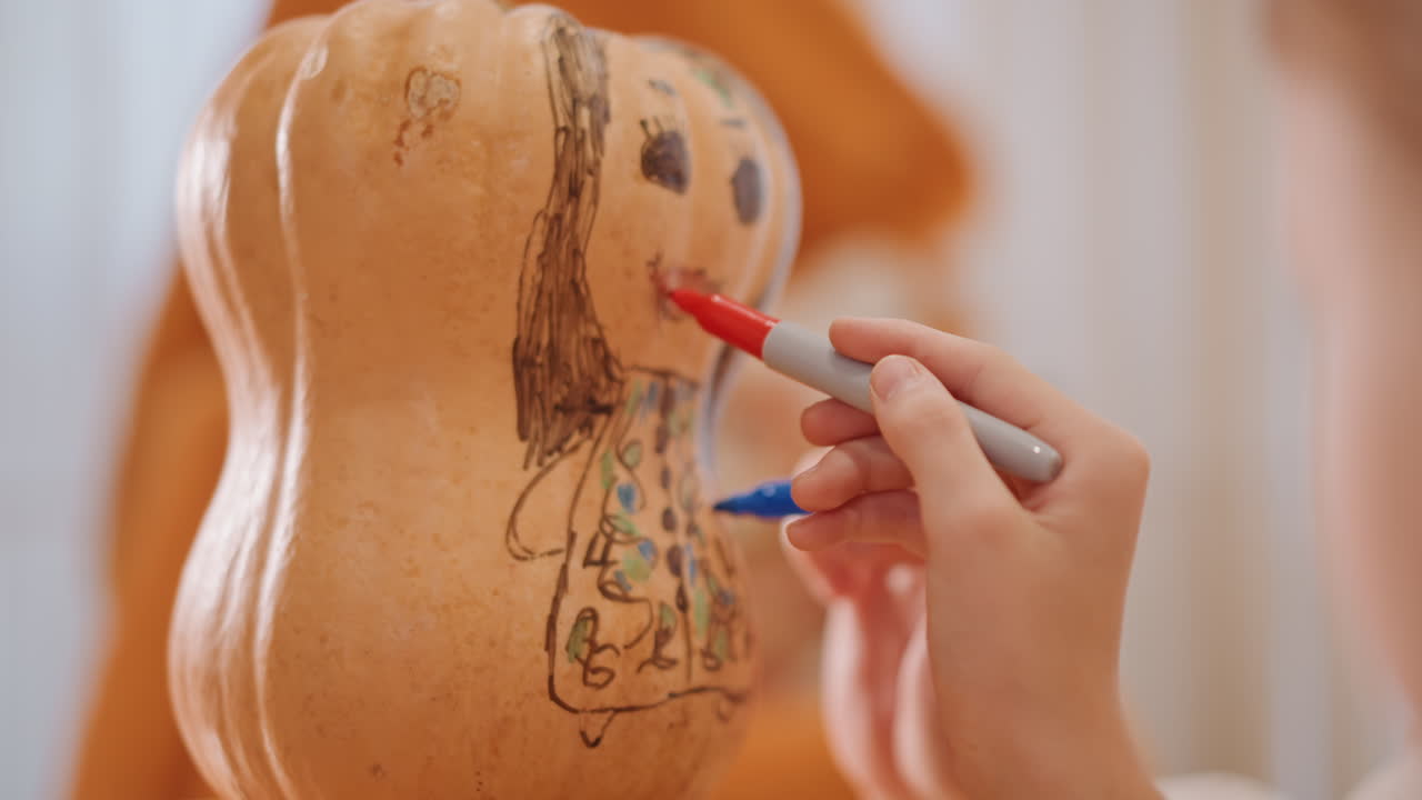 Bambino che decora una zucca con i pennarelli, primo piano di piccole mani che disegnano una faccina sorridente e un vestito sul frutto. Calda luce autunnale, scena accogliente in cucina, decorazioni natalizie fatte in casa per la stagione della torta di zucca.