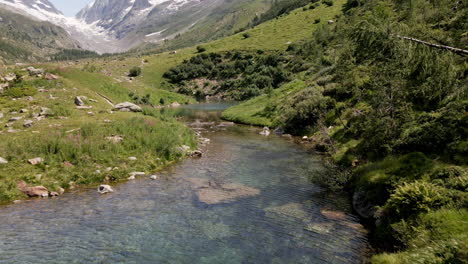 aerial view advancing through a mountain lake of turquoise clear water and following a river in the summer, with snowy mountains in the background