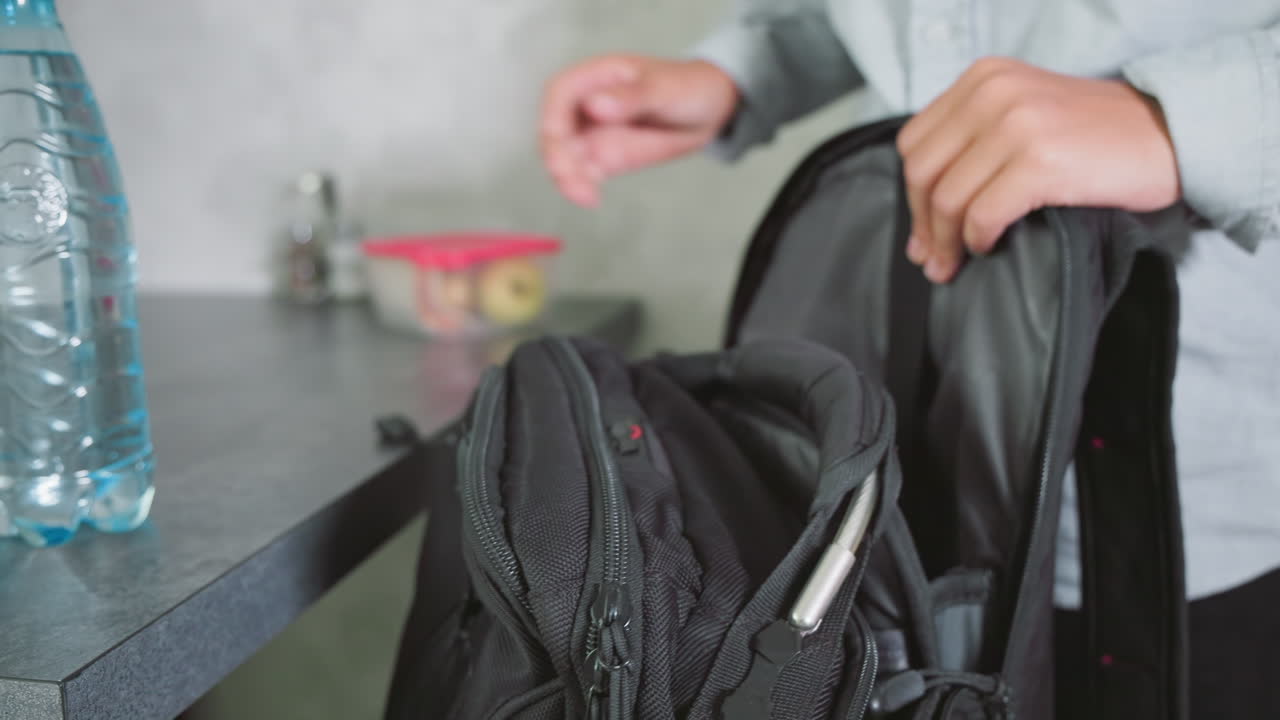 boy in casual shirt packing green notebook into black school backpack on kitchen counter with water bottles, lunchbox, and books, preparing supplies for school day with focus and neat arrangement