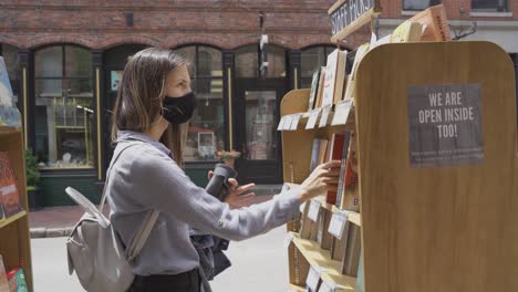 Frau-Mit-Maske,-Die-Bücher-An-Einem-Sonnigen-Tag-In-Einer-Buchhandlung-Im-Freien-In-Portland-Auswählt