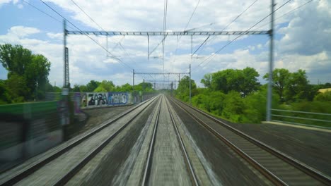 pov fast moving train with three tracks view traveling to a different stations with electrical cables above graffiti walls on the sides with trees forests fields and partly cloudy freedom travel