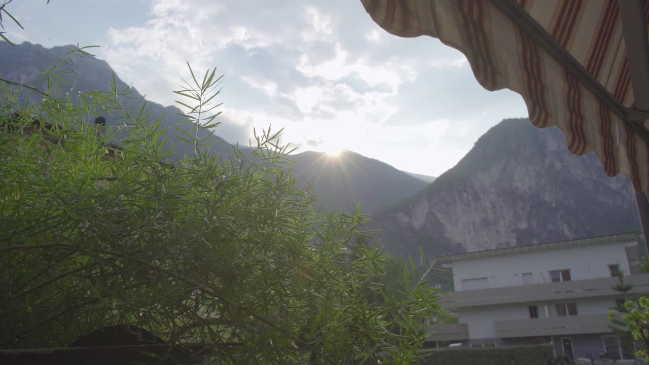 Sun is shining bright behind the top of the italian alps in Riva Del Garda with green plants in the foreground, Beautiful balkon view with mountainscape