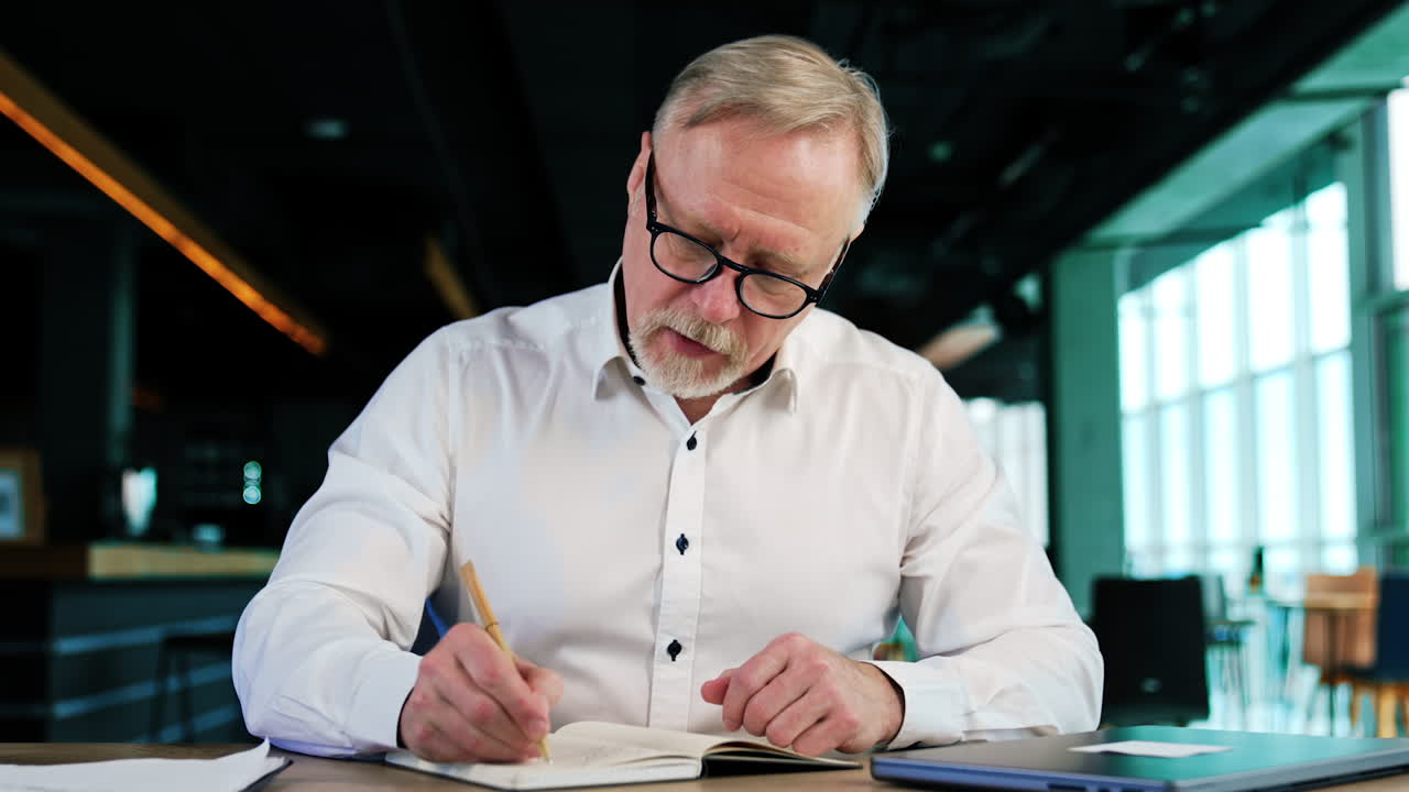 Focused Caucasian man in glasses writing in his paper notebook. Busy man taking notes at work. Close up.