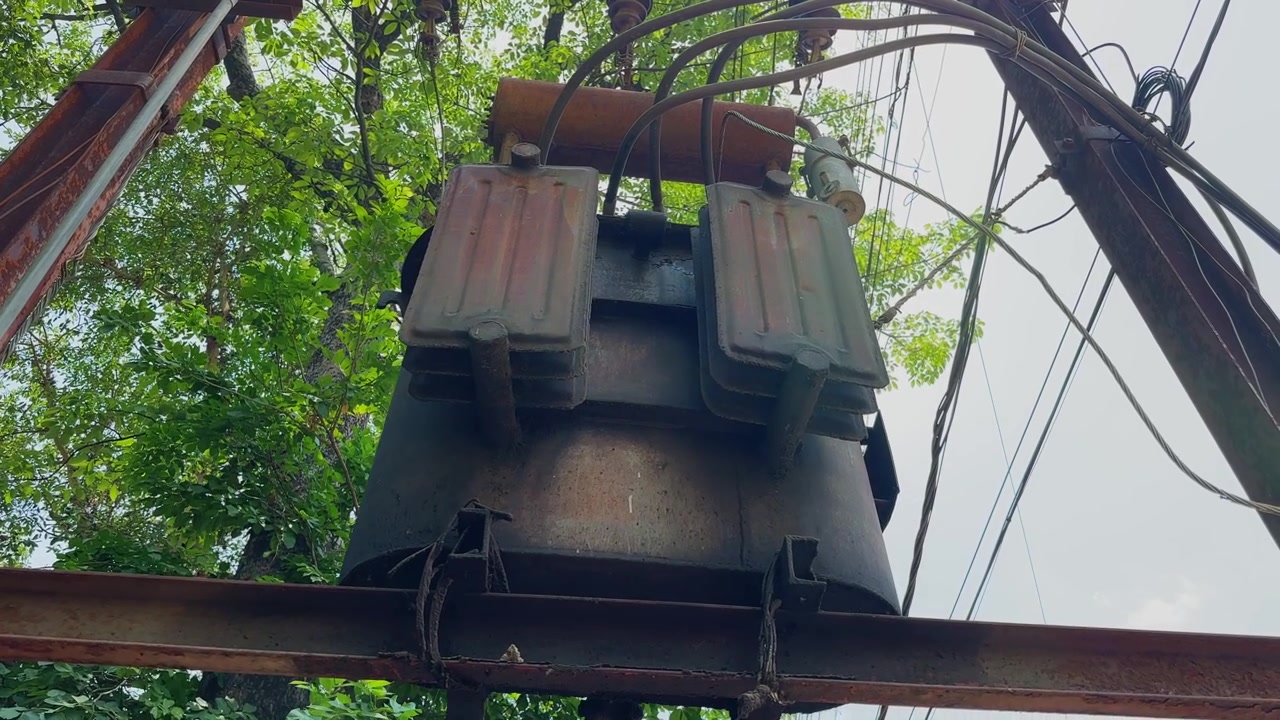 Telecamera che orbita attorno a un trasformatore di distribuzione elettrica montato su palo, catturando cavi elettrici, isolatori e infrastrutture di servizio incorniciati dagli alberi circostanti e dal cielo diurno