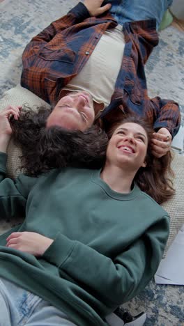 Vertical-video-of-a-happy-brunette-girl-in-a-green-jacket-lies-head-to-head-with-a-guy-in-a-checkered-shirt-on-the-floor-and-smiles