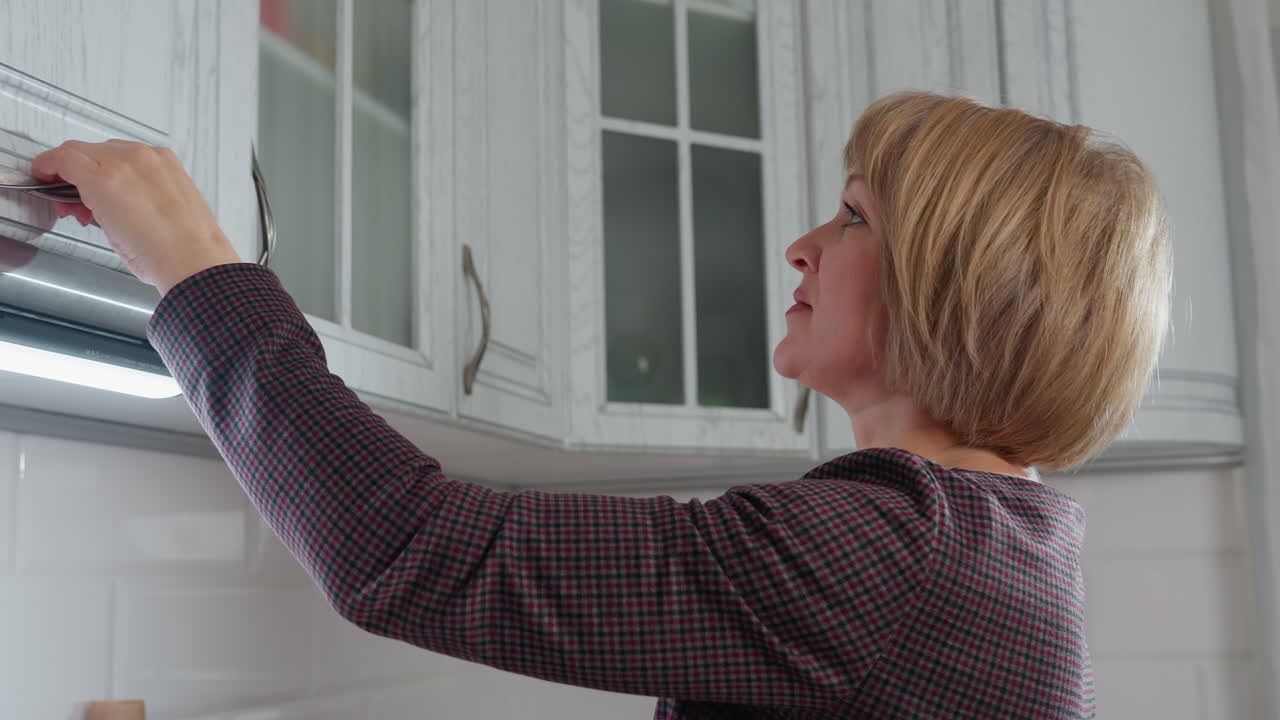 Huisvrouw die een item terugplaatst in een bovenkastje in de keuken terwijl ze het sluit, gekleed in een geruite outfit, verlicht door onderkastverlichting, moderne witte keuken