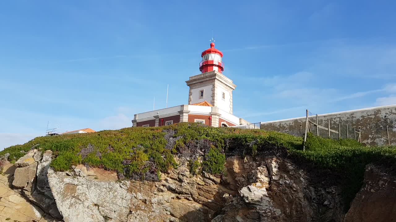 Red-domed Ponta do Pargo Lighthouse stands on layered cliff above Atlantic sea, surrounded by lush coastal flora and green grass, iconic western viewpoint on Madeira island under clear blue sky
