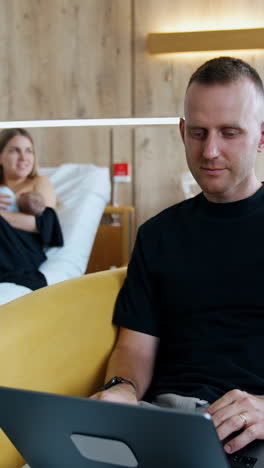 busy man is focused on his laptop sitting in the armchair. woman at backdrop feeding her baby lying in bed. vertical video