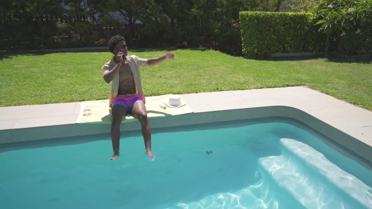 Man in swim trunks answering incoming call on phone, gesturing at yard and kicking feet poolside