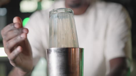 bartender holding cocktail shaker, carefully placing lid to seal it, preparing drink in bar. behind, illuminated liquor shelves and heineken neon sign glowing brightly, enhancing atmosphere