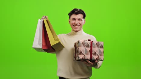 el hombre está mirando a la cámara y sonriendo. está sosteniendo un regalo y recogiendo bolsas de compras. está de pie sobre un fondo verde. pantalla verde. 4k.