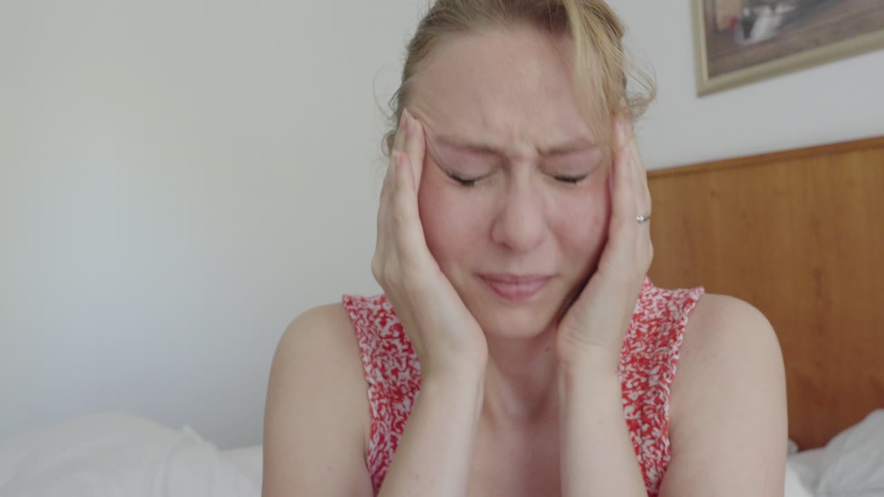Close-up of a woman pressing her temples with both hands, eyes closed in pain. Facial expression shows severe migraine or stress-related headache indoors