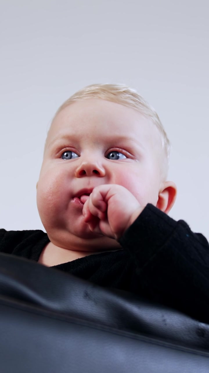 Small blond baby boy with plump cheeks is on the black leather sofa. Low angle view on the child of eight months old. Vertical video