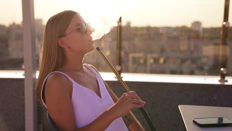 blonde lady in white dress smoking hookah sitting on the building roof. woman in sunglasses enjoys shisha looking at the cityscape at sundown.