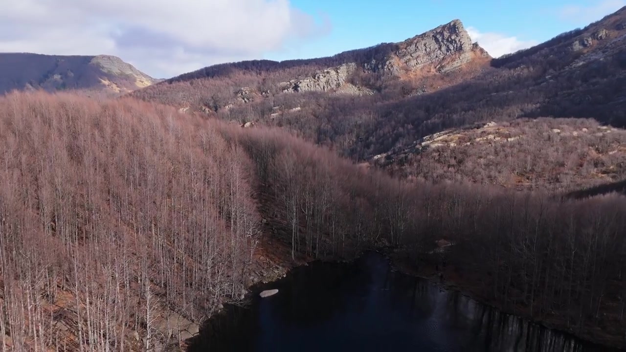 A cinematic aerial shot of the Lagoni glacial lake in the Apennines, featuring bare winter trees and rugged mountain peaks under a blue sky. Ideal for nature and travel documentaries