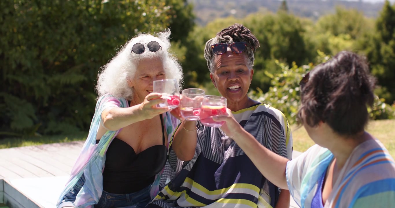 Tre donne in costume da bagno alzano bicchieri senza stelo, brindando e sorseggiando bevande rosa a bordo piscina.