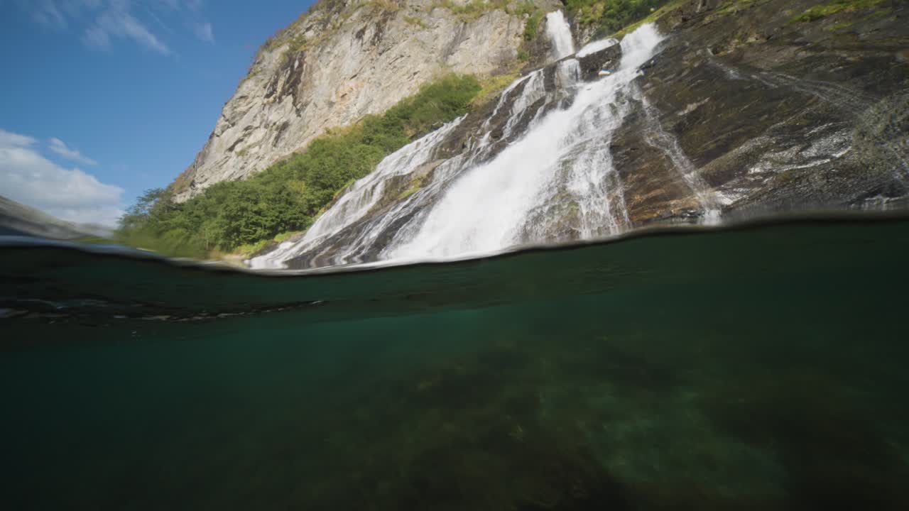 A close-up over-under view of a cascading waterfall flowing into the calm waters of Geiranger Fjord, surrounded by lush greenery.