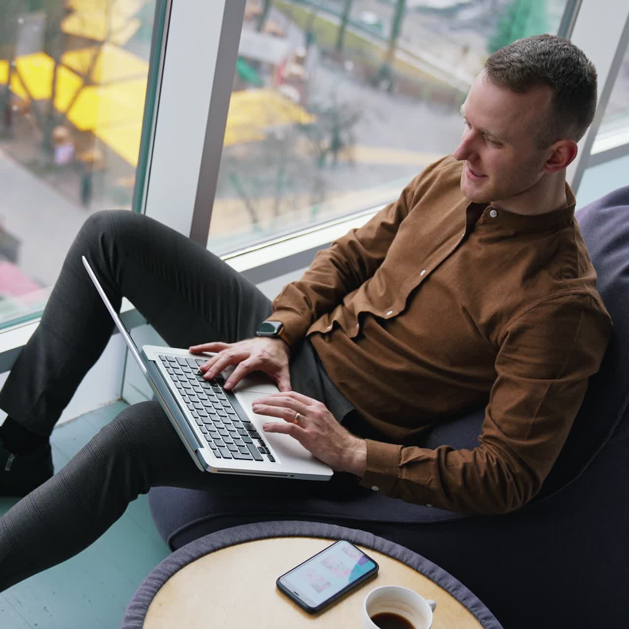 Young freelancer uses laptop. Handsome guy is sitting by the window with city view background and working on business with a wireless computer
