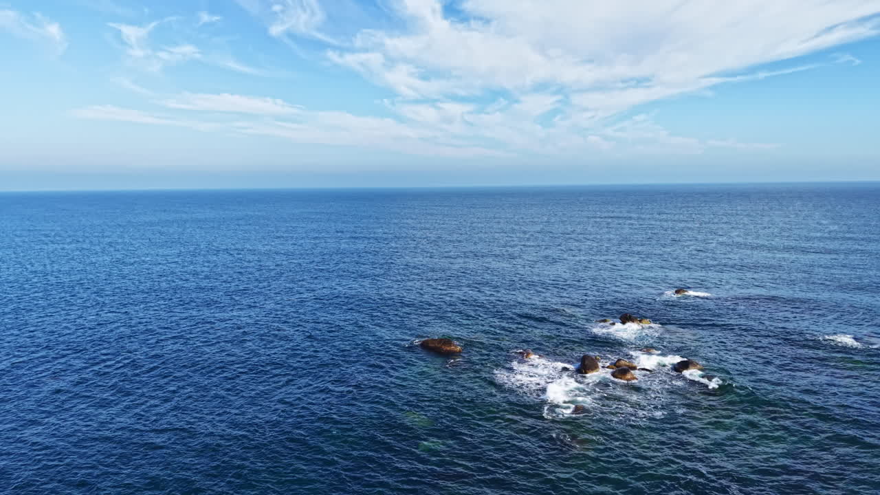 Vast blue ocean stretches to the horizon under a brilliant sky filled with wispy clouds near rocky coastline on a sunny day