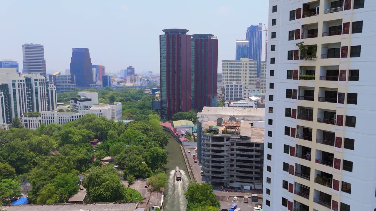 Vista de drone de Khlong Saen Saep em Asoke mostrando barco de canal movendo-se pela via navegável, vegetação ao longo das margens e arranha-céus circundantes em um distrito urbano denso.
