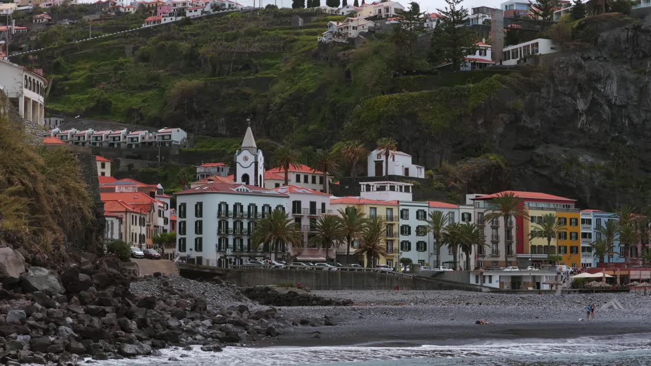 Charming coastal village with colorful houses, a church, and palm trees, nestled on a rocky beach with hills in the background