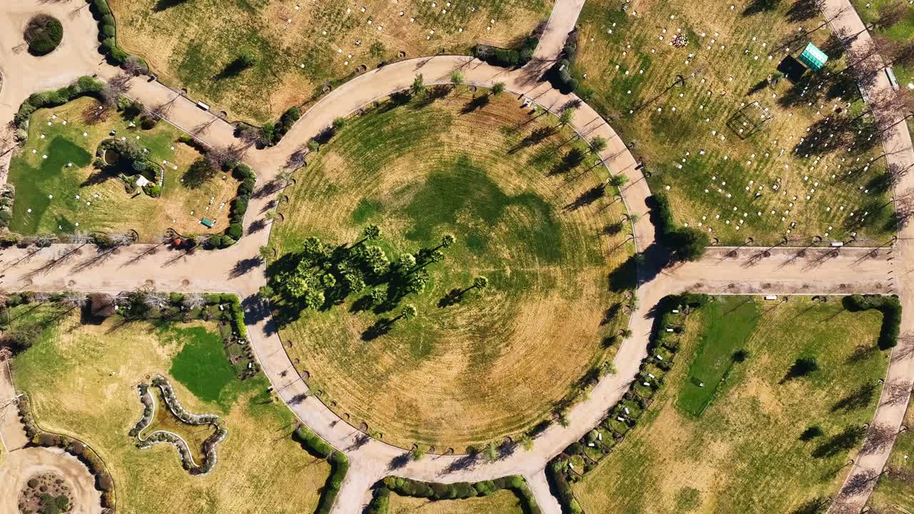Ascending top-down drone view of circular park with geometric pathways, dry grass, shadows, and landscaped urban green space, Santiago