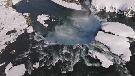 waterfall in winter landscape of iceland, revealing drone shot, flowing water, snow and rainbow above canyon