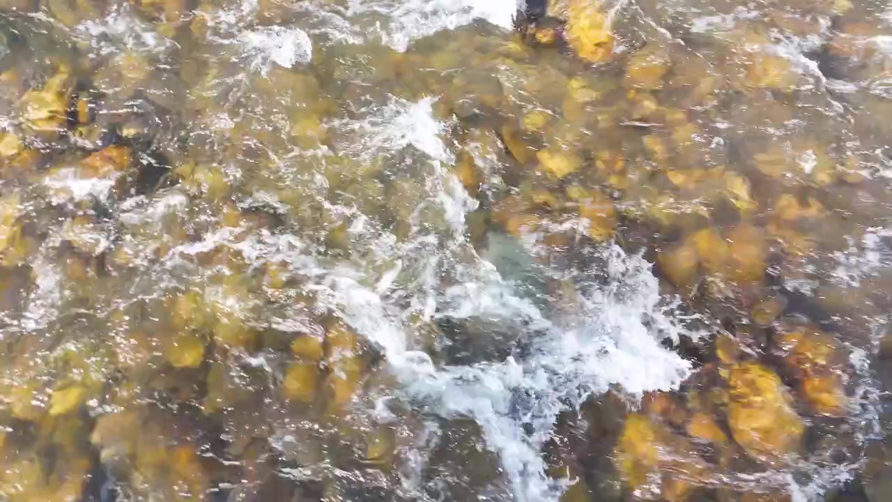 High angle shot of fresh river water rushing over smooth pebbles and submerged rocks.