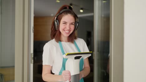retrato de una feliz dama de limpieza morena con auriculares inalámbricos negros y una camiseta blanca y un delantal azul que posa con una aspiradora de ventana en sus manos cerca de una puerta de vidrio en un apartamento moderno durante la limpieza