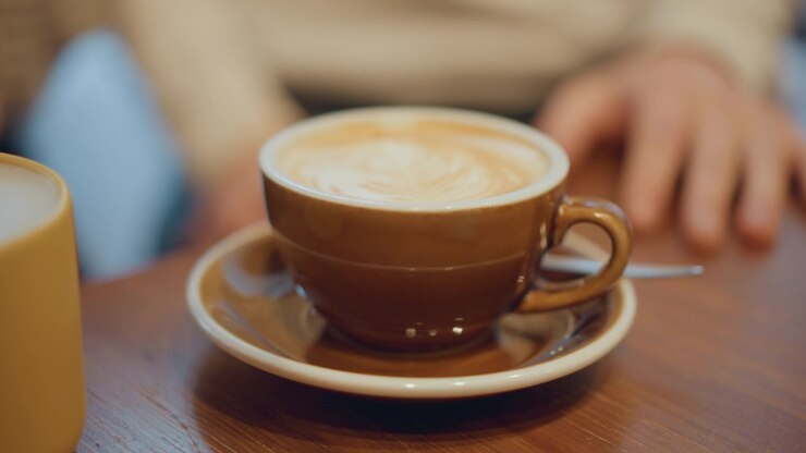 Primo piano di un latte macchiato in tazza di ceramica marrone su piattino, con una tazza gialla accanto. Una mano è appoggiata vicino al tavolo; l'illuminazione soffusa crea un'atmosfera calda. Ambiente interno con piano del tavolo in legno e sfondo sfocato.