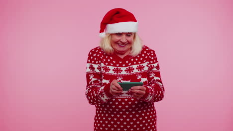 worried grandmother in christmas sweater enthusiastically playing racing video games on mobile phone