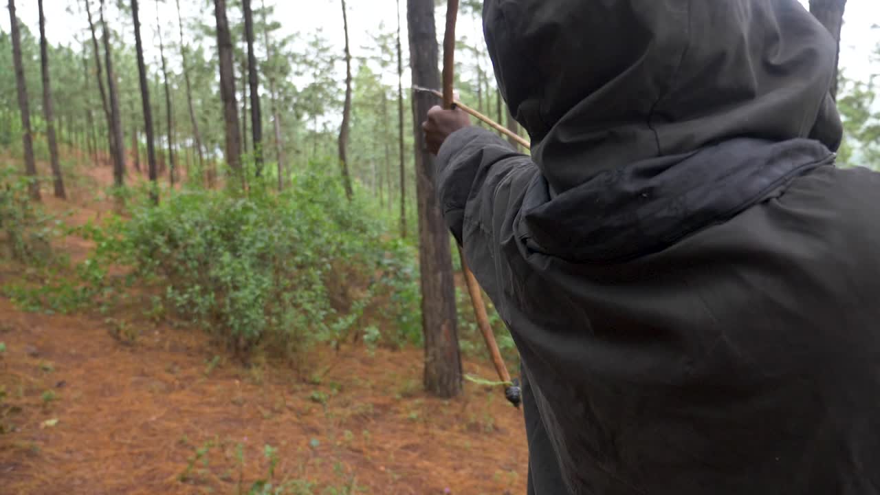 뒤에서 활과 화살로 조준하고 나무를 쏘는 후드와 은폐를 한 아프리카 사냥꾼의 슈퍼 슬로우 모션 샷
