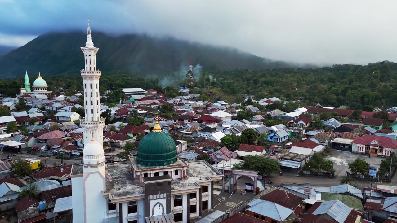 Masjid agung dikelilingi desa pegunungan padat penduduk dengan asap mengepul dan puncak tertutup awan di Sembalun Lombok, rekaman drone udara.