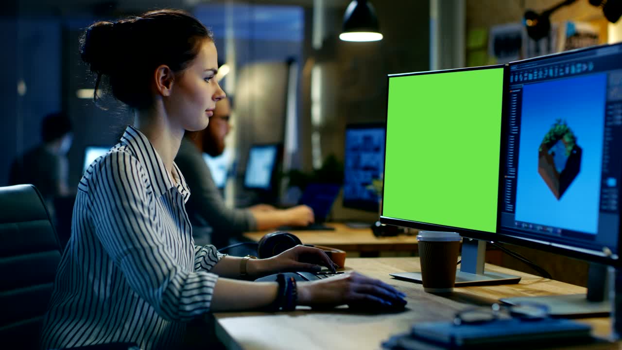 una hermosa mujer profesional creativa trabaja en una computadora personal con una pantalla verde de maqueta, la segunda pantalla muestra gráficos de juego en 3d. ella trabaja en un estudio creativo.