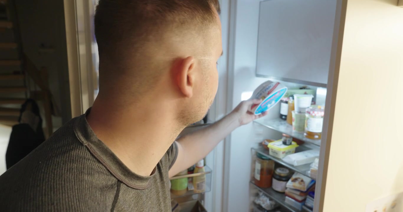 Muž se dívá do otevřené lednice a hledá jídlo. Koncept každodenního domácího životního stylu: hlad, prázdná lednice, potraviny a rozhodování.