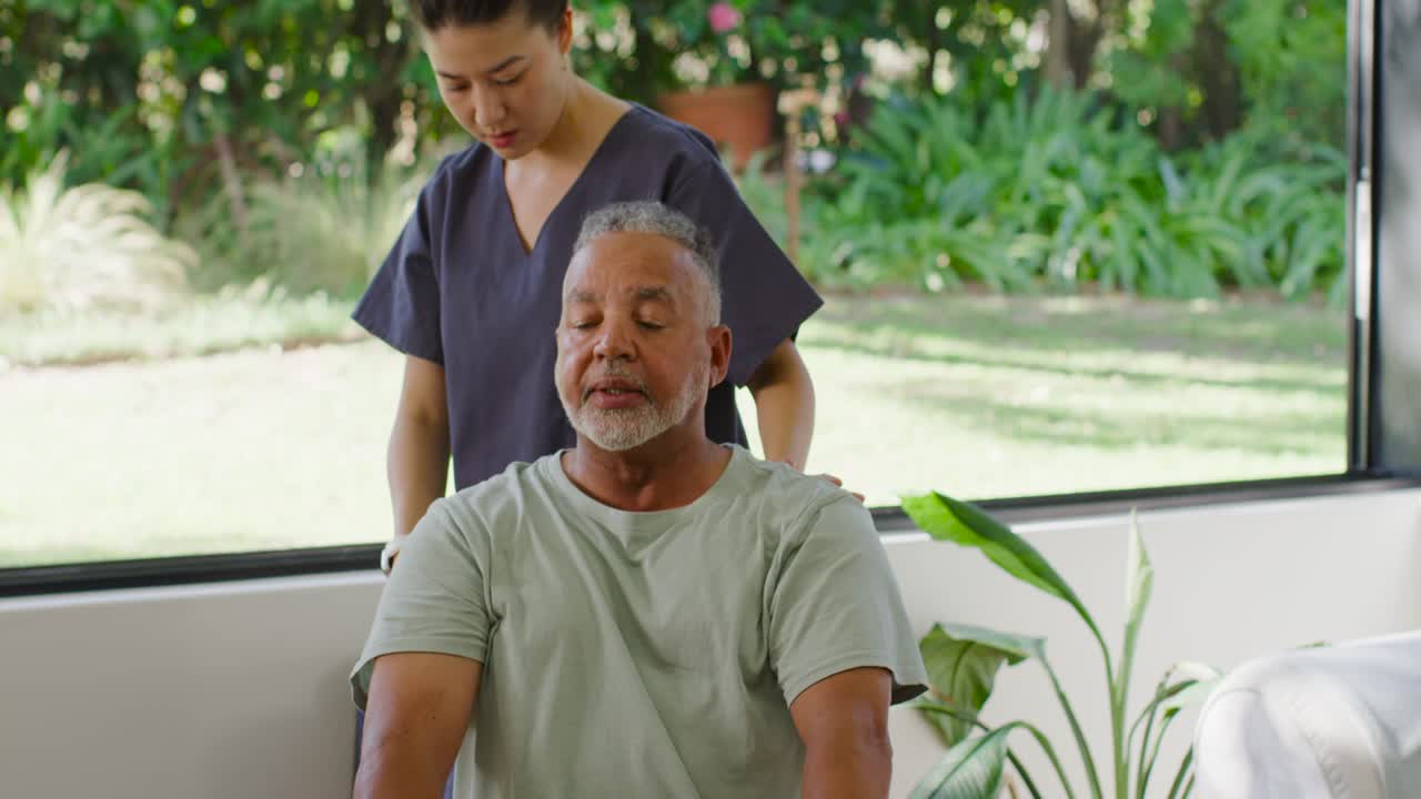Woman caregiver in scrubs placing hand guiding senior male for rising near garden window with watch
