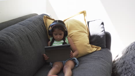 biracial girl with headphones uses a tablet on a gray couch