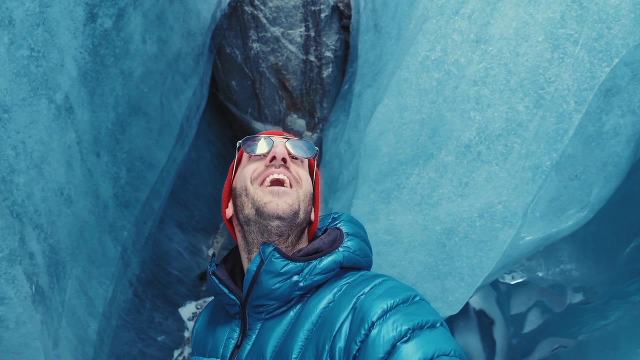 Hiker in a red beanie and blue jacket films himself inside a vivid blue Morteratsch Glacier ice cave, looking around in awe.