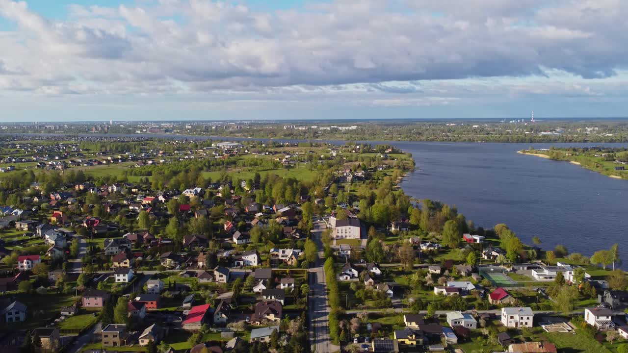 Slow drone pan over Katlakalns residential area, showing houses, Daugava river shoreline, tree lines, and Riga skyline on the horizon, suburban expansion, scattered clouds and soft springtime light