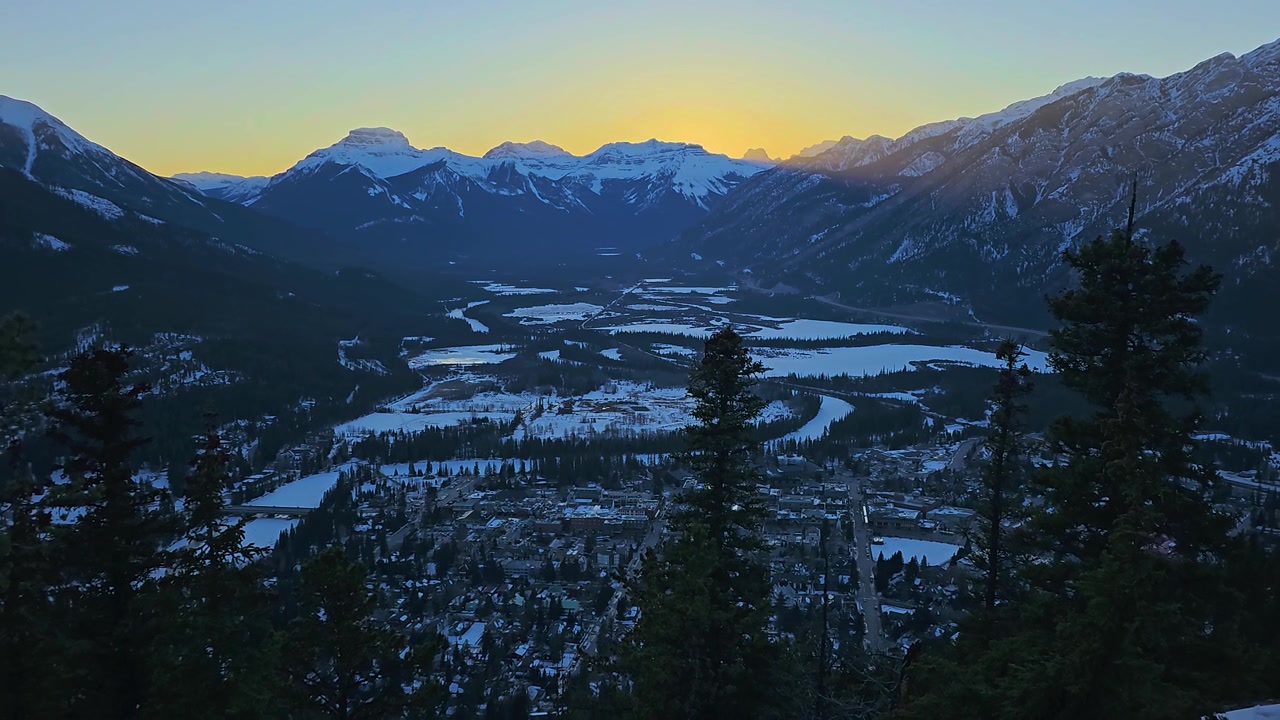 Cảnh hoàng hôn vàng rực trên Banff, Canada, với những ngọn núi phủ tuyết, rừng thông, thung lũng đóng băng, các tòa nhà thị trấn, bầu trời rực rỡ, một điểm đến tuyệt đẹp ở Dãy núi Rocky và du lịch mùa đông.