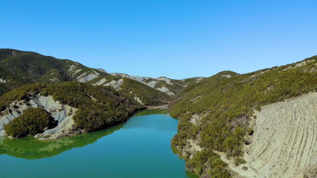 Lake with calm clear water reflecting mountains with green vegetation under bright blue sky background