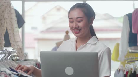 close up of asian female owner of clothing store enjoys using a laptop to check stock on rails