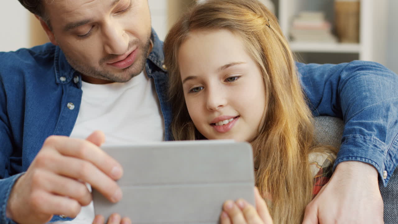 태블릿 컴퓨터에서 무언가를 보는 동안 소파에 앉아 예쁜 십대 딸을 껴안고 있는 잘생긴 아버지를 닫습니다